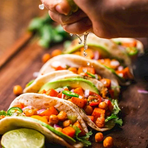A hand squeezes lime over tacos filled with chickpeas, avocado, and vegetables on a wooden board, garnished with some lime slices.