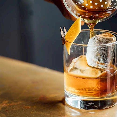 A beverage being poured from a strainer into a glass with ice and a citrus peel garnish, placed on a polished surface.